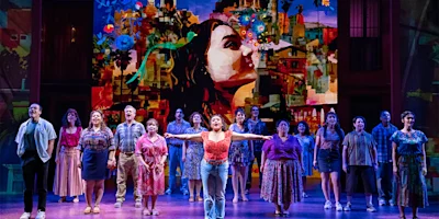 A large ensemble cast performs on stage in front of a colorful mural depicting a woman's face and a cityscape, with one performer standing front and center with arms outstretched.