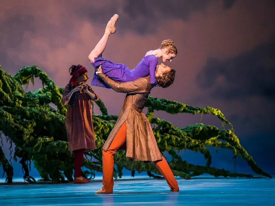 Production photo of The Winter's Tale, showing ballet dancers performing a lift on stage; a dancer in blue is elevated by another in brown. A person in a red outfit stands in the background beside green, vine-like decor.