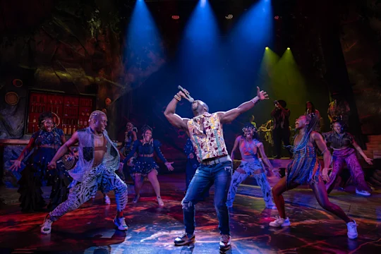 A man sings passionately into a microphone on stage, surrounded by dancers in colorful costumes under dramatic lighting.