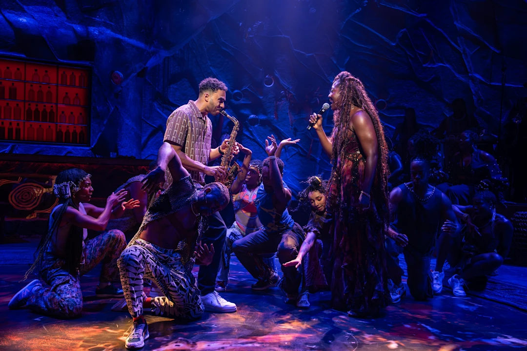 A group of performers in colorful costumes act and sing on a dimly lit stage decorated with dramatic lighting and set pieces.