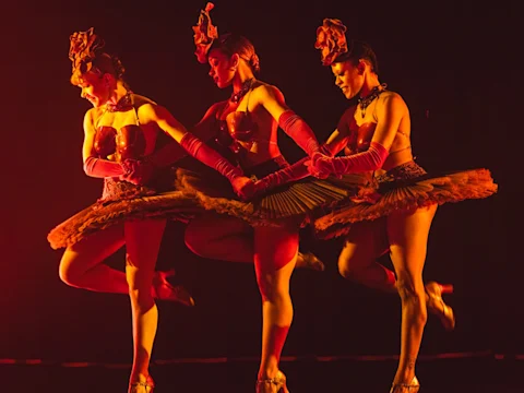 Three female dancers in red costumes and headpieces perform on stage, holding hands and lifting one leg, illuminated by dramatic orange-red lighting.