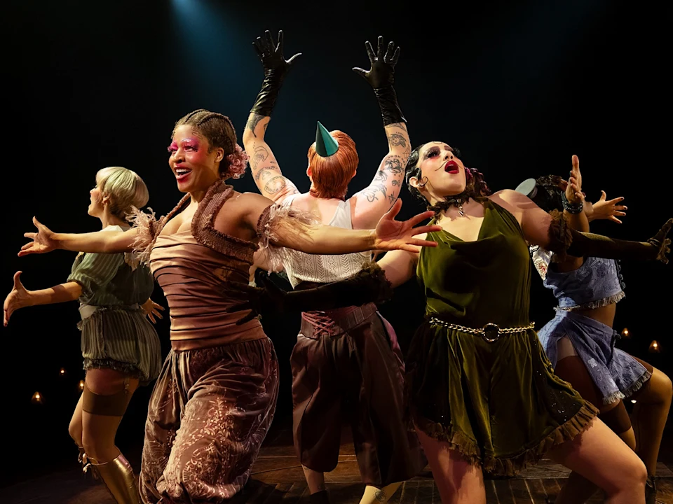 Six performers in colorful costumes stand on stage with dramatic poses and expressive faces, illuminated by stage lights against a dark background.