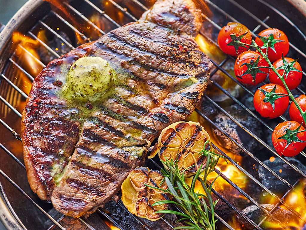A thick-cut steak on a barbecue with tomatoes and garlic.